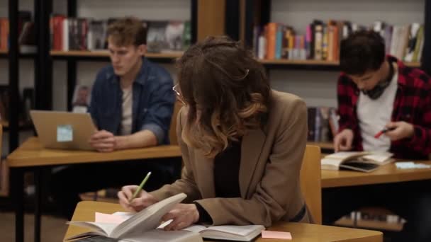 Chica europea rizada con chaqueta marrón sentada en la biblioteca de la universidad y trabajando en su tesis, preparándose para los exámenes, luego mira una cámara y sonríe. Sus compañeros de clase y estantes de libros en el fondo — Vídeos de Stock