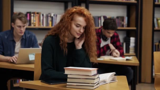 Retrato de uma jovem estudante encaracolada de cabeça vermelha trabalhando em sua tese ou se preparando para o exame. A ler um livro sentado à mesa na biblioteca. A sorrir para a câmara. Colegas do sexo masculino em segundo plano  - — Vídeo de Stock