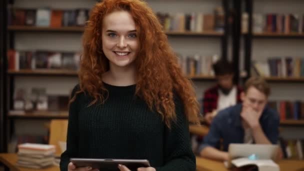 一个漂亮的长长的红头发的欧洲女生站在高中图书馆里，面带微笑地看着相机，面带微笑。 教育、文学和人的概念。 同学们都很模糊 — 图库视频影像