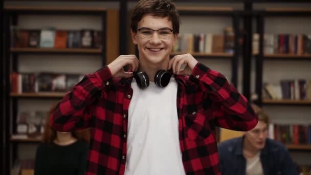 Gros plan portrait joyeux jeune homme riant jouissant d'une réussite de style de vie. Homme européen en chemise à carreaux et écouteurs à la bibliothèque. Bibliothèque et camarades de classe sur fond collège — Video