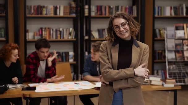 Portret van een aantrekkelijke kortharige Europese studente met een bril en een bruin jasje in de bibliotheek van de middelbare school, glimlachend kijkend naar de camera en glimlachend. Onderwijs, literatuur en mensen — Stockvideo