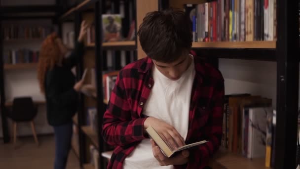 Fiatal jóképű férfi áll támaszkodva olvas egy könyvet - ingázó, diák, tudás koncepció. Egy kockás inges fiatalember könyvet olvas a főiskolai könyvtárban. — Stock videók