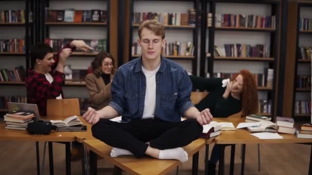 Káosz van a könyvtárban a tanulás alatt. Fiatal férfi diák meditál az asztalon a lótusz pózban, miközben az osztálytársai szórakoznak, papírt dobálnak a háttérbe — Stock videók