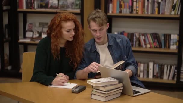 Piros hajú lány és jóképű srác, európai diákok együtt tanulnak, könyvet olvasnak a diákok könyvtárában, megvitatják és felkészülnek a vizsgákra. Üljön le az asztalhoz a könyvekkel, érzelmileg beszél. — Stock videók