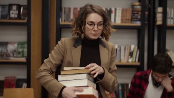 Slow motion footage of a girl in brown jacket and glasses walking by row with pile of books she took from the shelf. Other students sitting, studying in college, university library. Front view — 图库视频影像