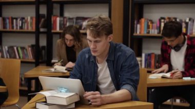 Blonde caucasian guy sitting in college library and working on laptop, preparing for exams then looks on a camera and smiling. His classmates and book shelves on the background