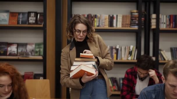 Spowolnione nagranie dziewczyny w brązowej kurtce i okularach chodzącej rzędem ze stosem książek, które wzięła z półki i zaczęła czytać książkę. Inni studenci siedzący, studiujący na studiach — Wideo stockowe
