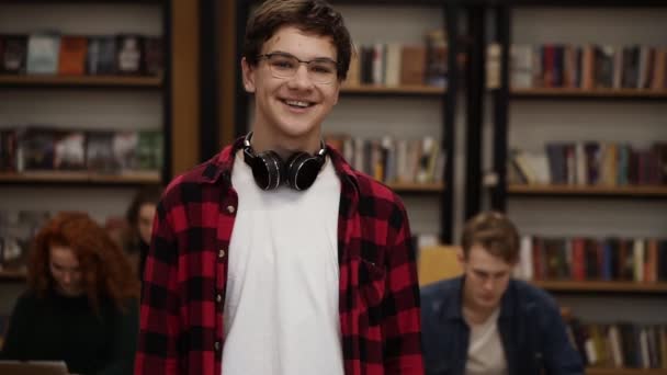 Portrait cheerful young man laughing enjoying successful lifestyle achievement, smiling to a camera. European male in plaid shirt and headphones in library. Bookshelf and classmates on background — 비디오