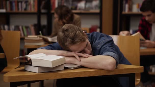 Joven estudiante europeo en una biblioteca universitaria o clase durmiendo en el escritorio con un montón de libros en él. Compañeros de clase y estantería en el fondo — Vídeo de stock