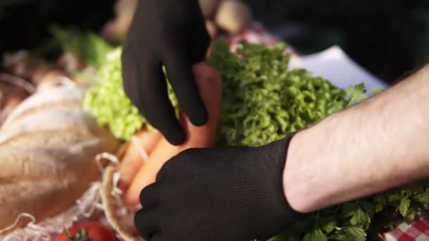 Primo piano delle mani maschili contadine con guanti neri che organizzano cibo biologico - insalata, carote biologiche a forma perfetta nel mercato agricolo in piedi all'interno in serra. Persone, raccolta e shopping concept — Video Stock