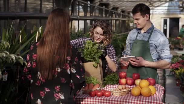 彼女の夫との陽気な販売の女性は笑顔販売野菜を話して温室市場の顧客に紙袋に新鮮なパセリを入れています。人々と健康的なライフスタイルのコンセプト — ストック動画