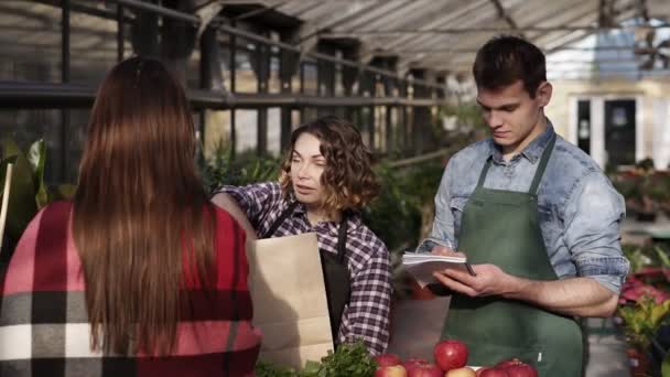 エプロンを身に着けているヨーロッパの販売員は温室で顧客に有機食糧を与えている。男はノートを作っている間女性は茶色の紙袋に緑、果物や野菜を梱包しています。人と健康 — ストック動画