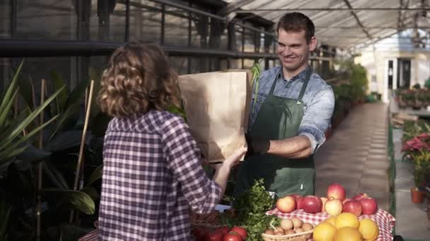 Potrait de cliente femenina contenta y sonriente que recibe una bolsa de papel con comestibles orgánicos. Caucásico, vendedor alto en delantal verde está vendiendo verduras en invernadero interior — Vídeo de stock