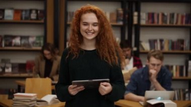 Portrait of an attractive long red haired european girl student standing in high school library smiling smiling looking at camera while holding a tablet. Education, literature and people concept