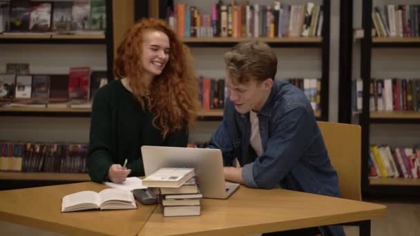 Front view of two young students female with long red hair dressed in black sweater and male in blue shirt talking in the library. Two students with laptop sitting side by side working on project — 图库视频影像