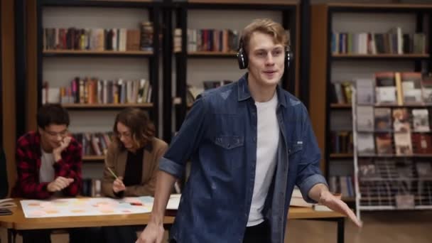 Красивый, взволнованный европеец в джинсовой рубашке исполняет выразительный танец, слушая любимую музыку в наушниках в академической библиотеке на книжных полках и фоне одноклассников. Медленное движение — стоковое видео