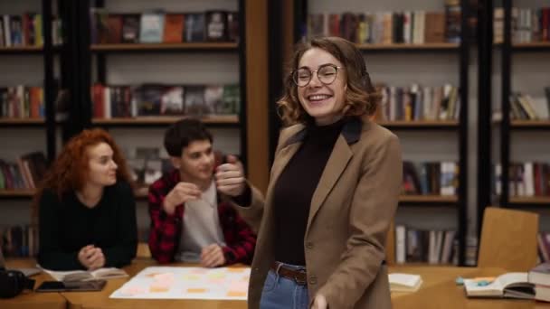 Retrato de alegre mujer europea excitada en chaqueta y jeans realizando danza expresiva mientras escucha música en auriculares en la biblioteca académica contra estanterías de fondo y compañeros de clase alrededor — Vídeos de Stock