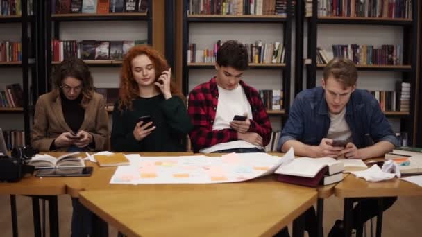 Groupe de quatre étudiants universitaires deux hommes, deux femmes assises à la table ensemble pendant la pause et à l'aide de smartphones textos, surfer sur Internet tout en étant assis dans la bibliothèque uni retour à la — Video