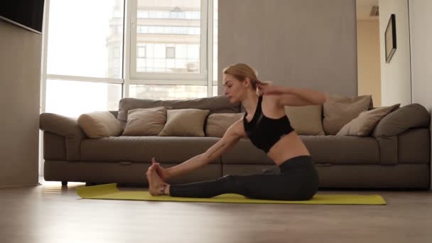 Blonde, caucasian woman in black leggings and sports bra doing youga or stretching body while sitting on a training mat at bright living room with a big couch on the background — Stock Video