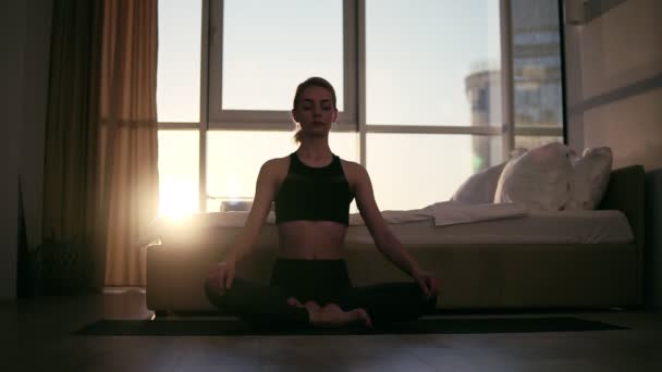 Splendida scena di donna adulta bionda che medita in posizione di loto a casa seduta sul tappetino yoga, godendo di esercizi. Giovani, stile di vita sano e concetto di interni. Lieve tramonto sullo sfondo — Video Stock