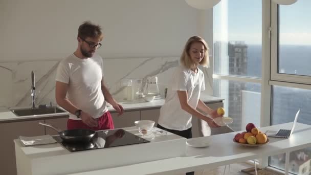 Jeune couple attrayant en vêtements domestiques sur une cuisine studip lumineux cuisiner le petit déjeuner simple.Bonne femme embrasser son mari, homme casser des œufs pour une omelette. Mouvement lent — Video