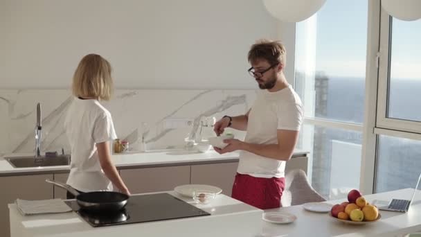 Relaks kaukaski mężczyzna przygotowuje śniadanie dla dwojga w kuchni w domu i mieszanie jajek w misce, blondynka przygotowuje przyprawy. Szczęśliwa para w białych koszulkach, uśmiechnięta i gadająca. Dzieciak ze studia. Promienie słońca — Wideo stockowe