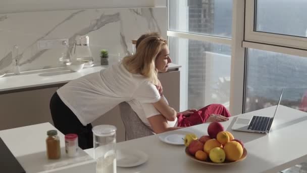 Sidovy över skäggig man som arbetar på bärbar dator sitter hemma på ett kök med panoramafönster. Ung fru omfamna honom bakifrån och bjuda in till frukost eller be om hjälp — Stockvideo