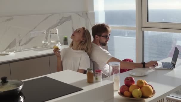 Side view of bearded man working on laptop computer sitting at home on a kitchen with panoramic windows. Young blonde wife sitting from behind and enjoying eating her apple in the morning — ストック動画