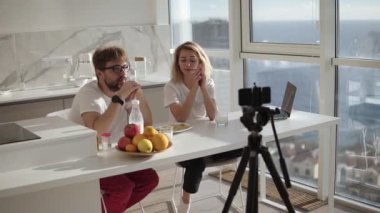 Genç mutlu çift blogcusu kameraya konuşuyor. Kahvaltı yaparken kamerada görüntülü konuşma kaydediliyor. Skype teknolojisi, neşeli insanlar, blog kameraları, online video...