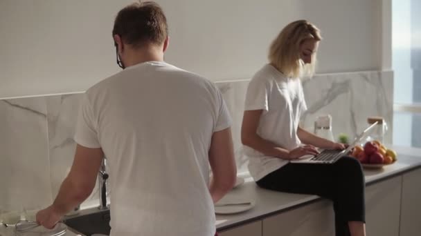 Paar verbringt Zeit miteinander im Gespräch in der Küche. Blonde Frau mit Laptop, auf einem Küchentisch sitzend, während ihr Mann das Geschirr spült. Morgengespräch. Zeitlupe — Stockvideo