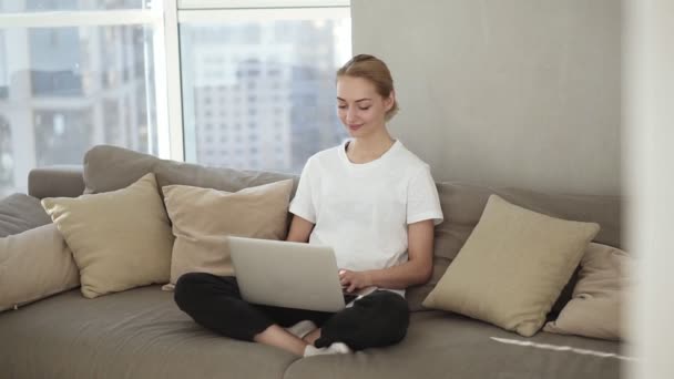 Mujer sonriente freelancer sentada en el sofá en ropa de hogar, freelance desde casa, escribiendo correo electrónico en el portátil. Enfocado chica rubia utilizando el ordenador mientras se sienta relajadamente en un sofá — Vídeo de stock