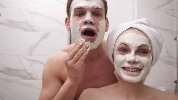 Feliz casal jovem positivo homem e mulher se divertindo enquanto estava em um banheiro após o chuveiro. Ambos brincando aplicando máscara branca no rosto. A divertir-me. Retrato casal juntos aplicando máscara facial — Vídeo de Stock