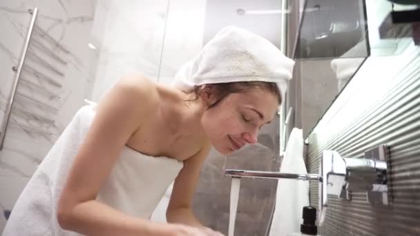 Una mujer parada en el baño cerca del lavabo con una toalla blanca en la cabeza y el cuerpo se lava la cara con agua del grifo. Mirando al reflejo del espejo y sonriendo. Vista de ángulo bajo — Vídeo de stock