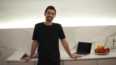 Mutfakta duran, gülümseyen, kameraya bakan iyimser, hayatının tadını çıkaran koyu kahverengi saçlı, kahverengi gözlü genç beyaz adamın portresi. Bir tezgahın üzerinde dizüstü bilgisayar