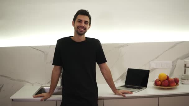 Portrait of young confident caucasian man standing in kitchen, smiling, looking at camera, full of optimism, enjoying his life, dark brown hair, brown eyes. Laptopn on a counter — Stock Video