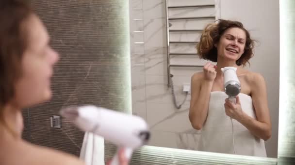 Mujer joven caucásica secándose el pelo corto en el baño, de pie en una gran toalla blanca y emocionalmente cantando secadora de usin como micrófono. Chica caucásica en estado de ánimo juguetón después de la ducha. Vista desde un — Vídeos de Stock