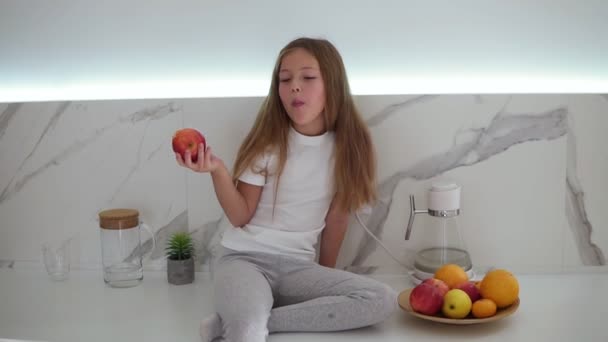Kislány hosszú, szőke hajjal, vörös almát eszik fényes, modern konyhában, miközben a pulton ül. Mellette egy tál friss gyümölcs. Lány élvezi friss alma — Stock videók