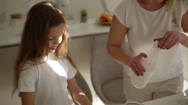 Mor hjälper och lär sin söta dotter i moderna mysiga vitt kök för att laga tårta eller pannkakor. Flicka tillsätta mjölk och blanda ingredienser. Lycklig familj. Förhållande mamma och dotter — Stockvideo