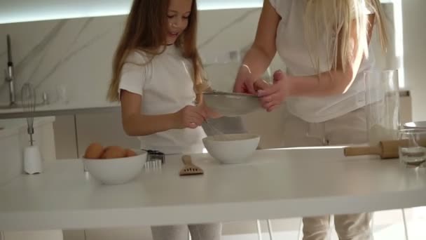 Madre ayudando y enseñando a su linda hija en la cocina blanca acogedora moderna para cocinar pastel o panqueques. Madre añadiendo harina a través de un tamiz y mezclando ingredientes. Divirtiéndose con harina. Movimiento lento — Vídeos de Stock