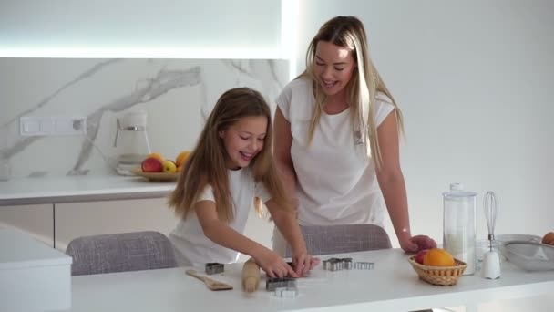 Concepto de familia, cocina y horneado - madre feliz y la pequeña hija cortan galletas en forma de mesa blanca en la cocina del hogar. Mamá e hija cocinando y divirtiéndose juntas - bailando, haciéndose cosquillas — Vídeos de Stock