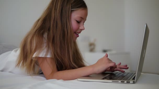 Dizüstü bilgisayarında yazı yazan uzun saçlı Avrupalı güzel bir kız. Gülümseyen küçük kız dizüstü bilgisayarı izle ve yatağa uzan. Yan görünüm, yavaş çekim — Stok video