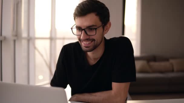 Портрет зубастого улыбающегося человека в очках с помощью ноутбука. Фрилансер работает дома за компьютером. Красивый мужчина активно печатает на ноутбуке, общаясь с друзьями в большом доме с панорамными окнами и — стоковое видео