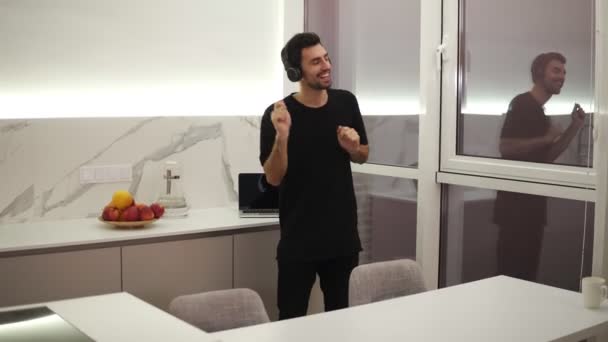 Hombre alegre en ropa negra está bailando en la cocina escuchando música con auriculares, divirtiéndose solo en apartamento moderno y ligero. Concepto de dispositivos y personas. Acelerado — Vídeos de Stock