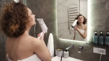 Neşeli, güzel genç bir kadın banyoda kısa saçlarını kurutuyor, büyük beyaz bir havluyla duruyor. Beyaz kız duştan sonra neşeli bir ruh hali içinde. Ayna yansımasından bak