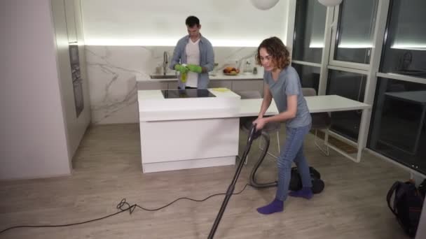 Putztag. junges Paar putzt seine Wohnung - Mann in Handschuhen wischt Küchenherd ab, Mädchen putzen Boden mit Staubsauger. saubere Studioküche abends zusammen. Zeitlupe — Stockvideo