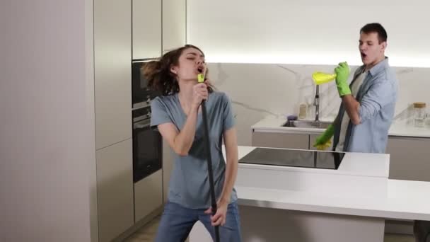 Deux jeunes dansant ensemble et chantant émotionnellement en utilisant un balai et des outils de nettoyage comme microphones pendant le nettoyage dans la cuisine. Couple heureux profiter du temps, se sentir comme des rock stars — Video