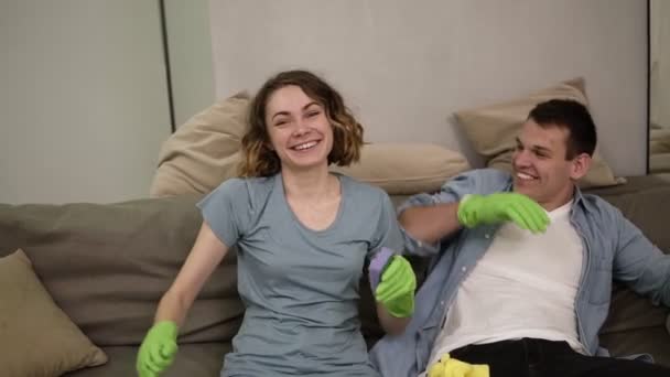 Mooie jonge vrouw en een man met groene rubberen handschoenen. De familie die samen het huis schoonmaakt. Gelukkig moe team. Huishouden huishoudelijk werk en schoonmaak. Zittend op een bank — Stockvideo