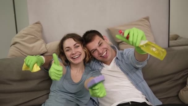 Retrato de um jovem e alegre casal em luvas de borracha verde. A família terminando a limpeza da casa juntos. Equipe cansada feliz, sentada em um sofá, olhando para a câmera, sorrindo, polegar para cima — Vídeo de Stock