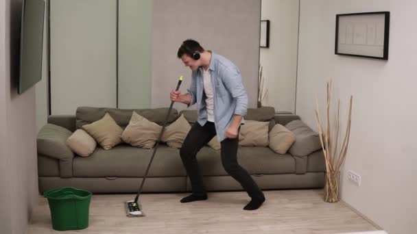 Ludzie, prace domowe i koncepcja sprzątania - szczęśliwy człowiek w słuchawkach z mopem sprzątanie podłogi i taniec w domu. Używając wacika jako gitary. Szara kanapa na tle — Wideo stockowe