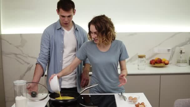 Glada par gör frukost tillsammans. Flicka häller blandade ägg från en skål till en varm kastrull på en köksspis. Maken står bakom och tittar. Långsamma rörelser — Stockvideo